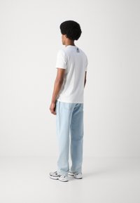Camiseta blanca de manga corta con un sutil logo negro en la parte posterior, combinada con jeans de mezclilla azul claro y zapatillas blancas con detalles.
