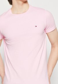 Mann, der ein hellrosa T-Shirt mit kurzen Ärmeln und einem kleinen rechteckigen Flaggenlogo auf der linken Brust trägt, vor einem schlichten Hintergrund.