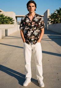 Ung mann står ute og har på seg en svart kortermet skjorte med blomster, hvite bukser og hvite joggesko, med hendene i lommer.