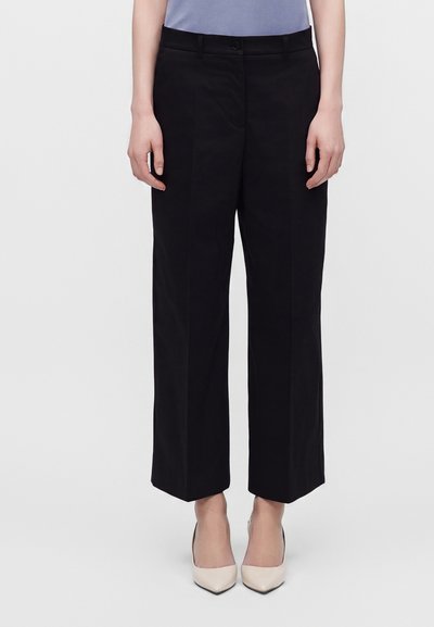 Femme portant un pantalon noir à jambes larges, un haut bleu clair et des chaussures beige à bout pointu, debout devant un fond uni.