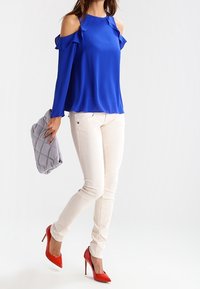 Kungablå kallaxelblus med volangdetaljer, kombinerad med vita smala jeans och röda high heels, hållande en grå vadderad väska.