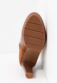 Botte cheville en daim marron, posée avec la semelle contre un mur blanc, montrant la semelle en caoutchouc texturée et le talon empilé.