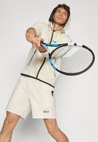Chaqueta deportiva color beige claro con capucha, detalles en negro en el cierre; pantalones cortos a juego, logo en los pantalones cortos; modelo sosteniendo una raqueta de tenis.