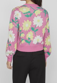 Frau in einem rosa Pullover mit großen weißen und gelben Blumen und grünen Blättern, kombiniert mit schwarzen Hosen, von hinten gezeigt.