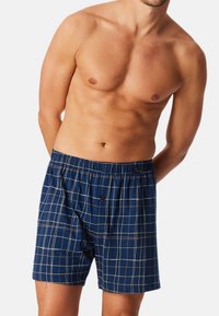 Pantaloni corti a scacchi blu navy realizzati in tessuto morbido, dotati di una vita elasticizzata e di un singolo bottone di chiusura. Motivo a scacchi con linee bianche e color caramello.