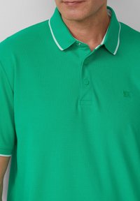 Groene polo met een gestructureerde stof, drie knopen bij de hals, en contrasterende witte accenten op de kraag en mouwen. Subtiele logodetail.