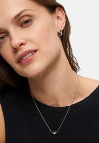 Silberne Kette mit einem kleinen Herzanhänger, getragen auf einem dunkel strukturierten Oberteil; das Model hat kurze Haare und minimales Makeup, wobei der Fokus auf dem Schmuck liegt.