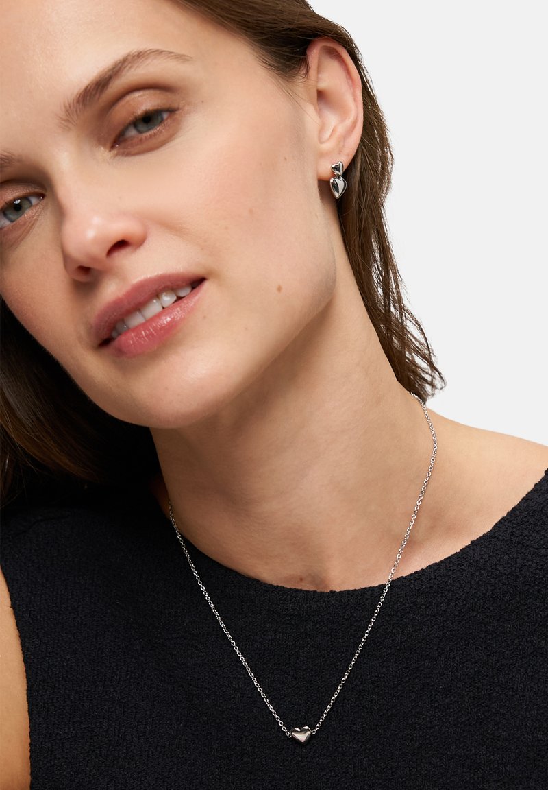 Silberne Kette mit einem kleinen Herzanhänger, getragen auf einem dunkel strukturierten Oberteil; das Model hat kurze Haare und minimales Makeup, wobei der Fokus auf dem Schmuck liegt.