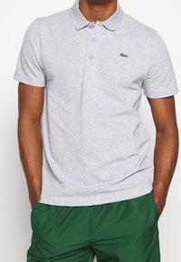 Man som bär en ljusgrå kortärmad polo t-shirt med en liten grön krokodil-logotyp och mörkgröna byxor, med händerna i fickorna.