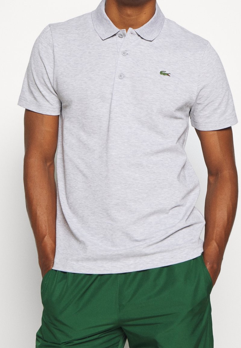 Man som bär en ljusgrå kortärmad polo t-shirt med en liten grön krokodil-logotyp och mörkgröna byxor, med händerna i fickorna.