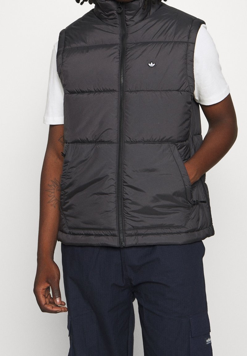 Černá vatovaná vesta s vysokým límcem, svislými švy, předním zipem a vyšívaným logem na levé straně hrudi. Má dvě boční kapsy.