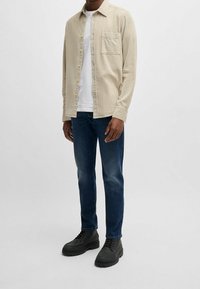 Man som bär en beige knapp-up skjorta över en vit t-shirt, mörkblå jeans och svarta snörskor, står på vit bakgrund.
