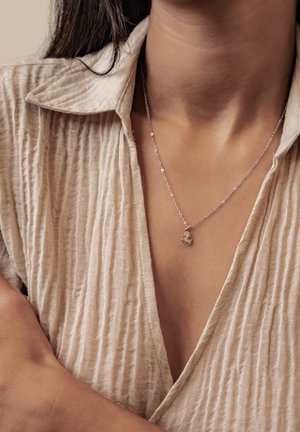 Frau trägt ein beiges, strukturiertes Hemd mit einer silbernen Halskette, an der ein kleiner Anhänger auf ihrer Brust ruht.