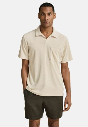 Man wearing a beige short-sleeve polo shirt with chest pocket and dark green shorts, standing with one hand in pocket against white background.