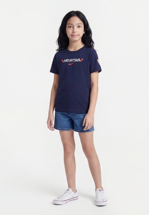 Fille aux longs cheveux foncés portant un t-shirt bleu marine « HRVATSKA », un short en jean et des baskets blanches, debout devant un fond blanc uni.