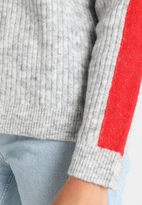 Maglione grigio a coste con un audace accento rosso sulla manica, realizzato in materiale morbido, con una vestibilità rilassata e una texture evidente.