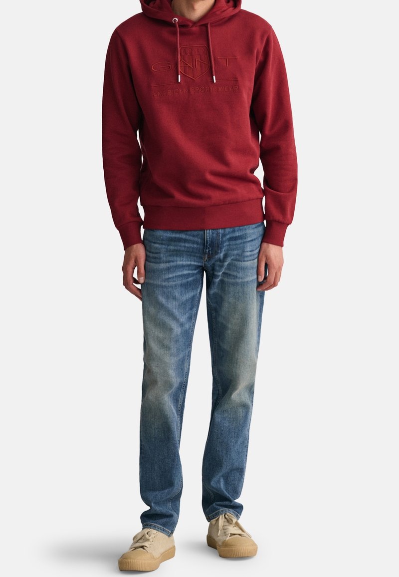 Man in een rode hoodie met logo, vervaagde blauwe jeans en beige sneakers, staand tegen een witte achtergrond.