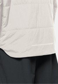 Bas du corps portant une doudoune beige clair sur un pantalon noir, montrant la texture du tissu de la doudoune et les coutures horizontales.