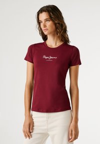 Žena s vlnitými hnedými vlasmi, oblečená v burgundskej tričku Pepe Jeans London a svetlobežových nohaviciach, stojí na jednoduchom svetlom pozadí.