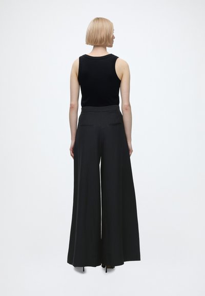 Femme aux cheveux blonds courts portant un haut noir sans manches et un pantalon noir à jambes larges, debout de dos sur un fond uni.