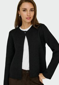 Schwarze, langärmelige Strickjacke mit rundem Halsausschnitt, offenem Vorderteil und einem Knopfverschluss. Weiche Textur, kombiniert mit einem weißen T-Shirt darunter.