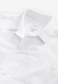 Bijela lanena košulja s okruglim ovratnikom na kopčanje, jednim džepom na prsima i dva bijela gumba sprijeda. Vidljiva etiketa s oznakom veličine 'M'.