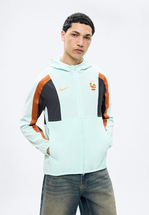 Joven con chaqueta Nike en azul claro, negro y naranja con el emblema de fútbol FFF y jeans azules desteñidos, de pie con las manos en los bolsillos.