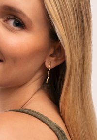 Złote skręcone kolczyki o falistym wzorze, wiszące z płatka ucha, na tle jasnej skóry i długich blond włosów.