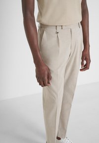 Pantaloni beige chiari su misura con tessuto strutturato, pieghe frontali e chiusura a bottone. Abbinati a un top a maglia neutra e sneaker bianche.