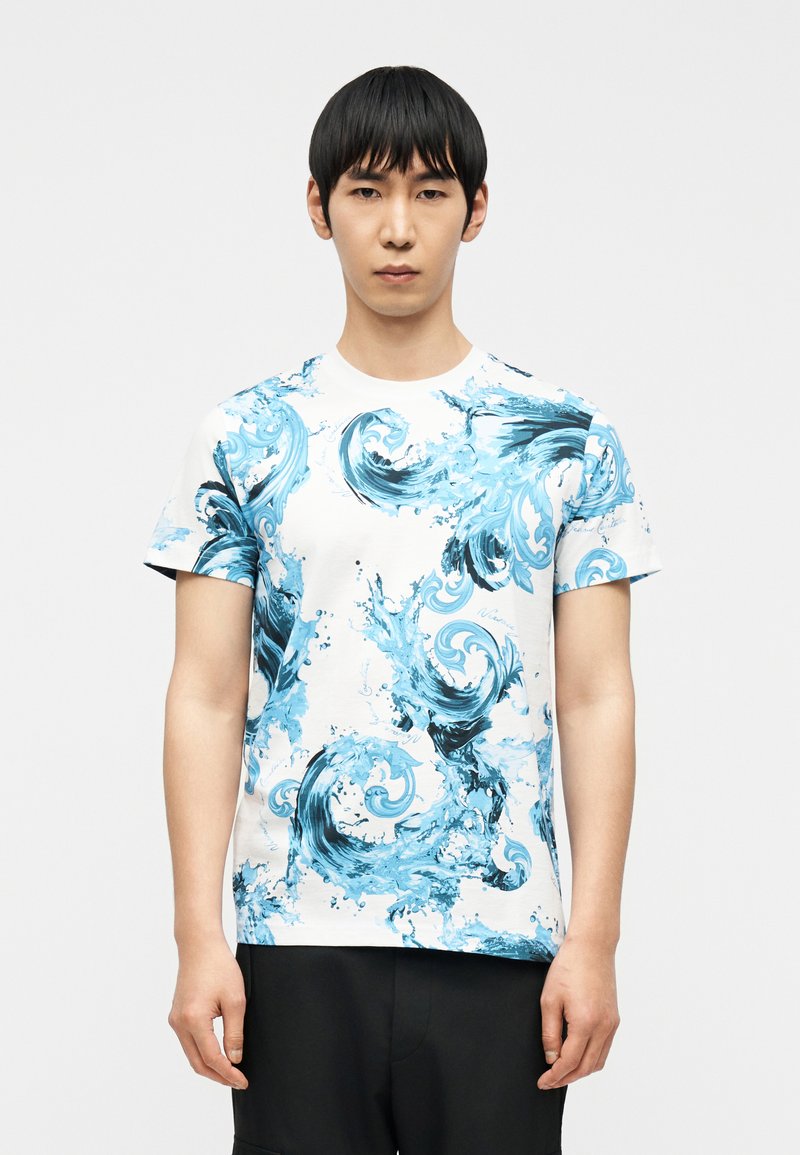 Άνδρας που φοράει λευκό κοντομάνικο μπλουζάκι με μπλε και μαύρα στροβιλιζόμενα μοτίβα κυμάτων νερού, στέκεται μπροστά σε μονόχρωμο φόντο.