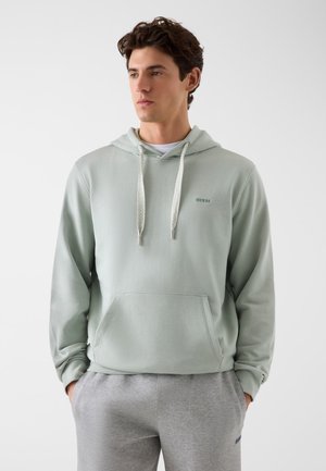 Jonge man die een lichtgroene capuchontrui met voorzak en grijze joggingbroek draagt, staand tegen een effen witte achtergrond.