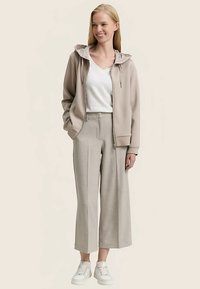 Felpa beige con cappuccio con zip e cappuccio a contrasto, abbinata a pantaloni ampi color grigio chiaro e sneakers bianche, caratterizzata da una vestibilità comoda e una texture morbida.