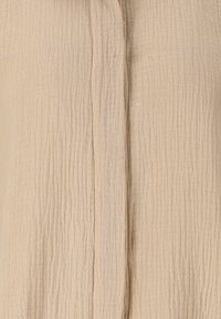 Beige textiel met een gekreukt patroon. Het materiaal lijkt lichtgewicht en ademend, met knoopdetails aan de voorkant.