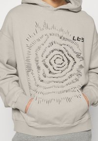 Sweat à capuche gris clair en tissu mélange de coton, avec un grand motif spirale noir sur le devant. Comprend une poche kangourou et des poignets côtelés.