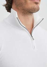 Pullover blanc zippé avec un col montant, texture de tissu lisse et un détail de fermeture éclair argentée au niveau du cou.