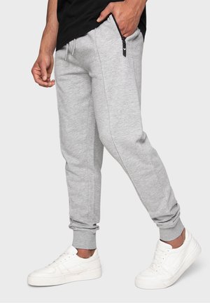 Homme portant un pantalon de jogging gris clair avec une poche zippée et des baskets blanches, debout devant un fond uni.