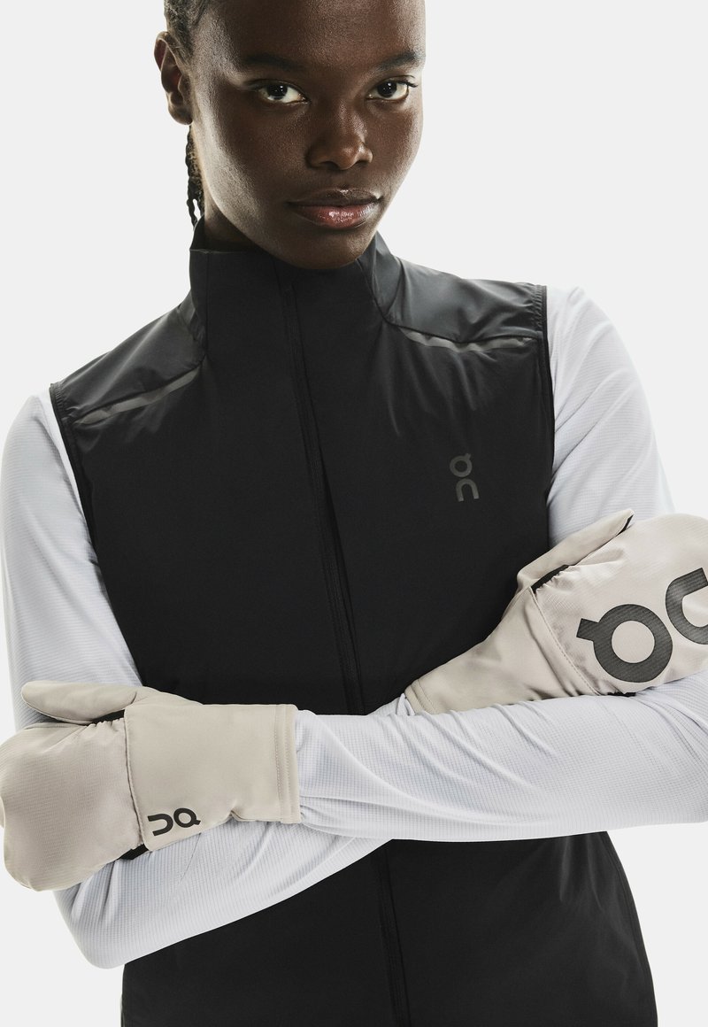 Gilet sans manches noir avec un col haut, superposé sur un haut à manches longues blanc. Des gants beige présentent un logo noir bien visible. Texture lisse.