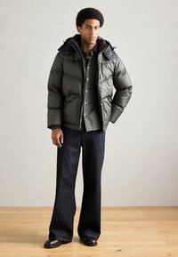 Jakna od pufova u tamno zelenoj boji s kapuljačom, s ukrasnim prošivom i bočnim džepovima. Kombinirana s prugastom košuljom i širokim tamnim trapericama.