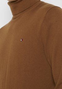 Brun rullekravebluse lavet af tekstureret strikket stof med et lille broderet logo i rød, hvid og blå på brystet.