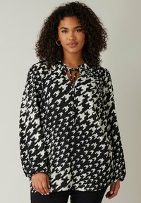 Bluse i sort/hvid houndstooth-mønster med lange ærmer, bindebånd ved halsen og løs pasform. Fremstillet af et let materiale.