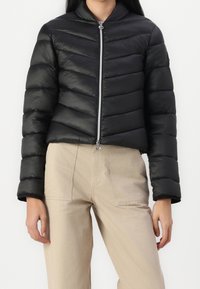 Czarna kurtka puchowa o skróconym kroju, pozioma pikowana faktura, wysoki kołnierz i przedni zamek błyskawiczny. Noszona z beżowymi spodniami.