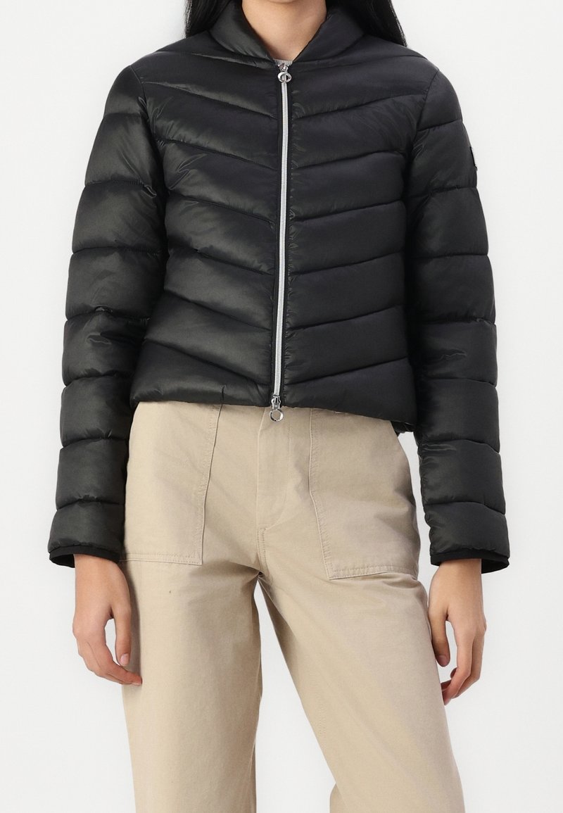 Czarna kurtka puchowa o skróconym kroju, pozioma pikowana faktura, wysoki kołnierz i przedni zamek błyskawiczny. Noszona z beżowymi spodniami.