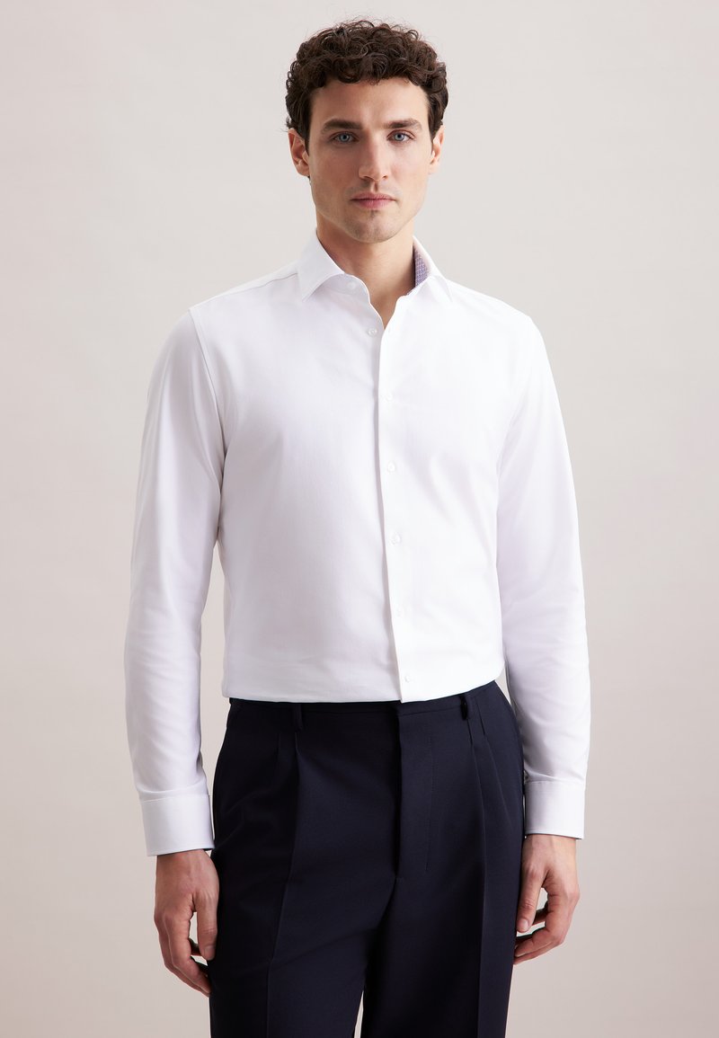Camicia bianca a maniche lunghe con colletto, realizzata in un tessuto morbido. Abbinata a pantaloni blu scuro. La camicia presenta una chiusura a bottoni e un design aderente.