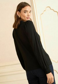 Fekete, hosszú ujjú felső, texturált felsőrésszel és ráncolt mandzsettákkal. A sima anyag karcsú, testhezálló siluettet biztosít, sötét farmerral párosítva.