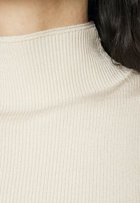 Maglione dolcevita beige chiaro con texture a coste e vestibilità aderente, caratterizzato da un collo semplice e pulito e da un tessuto morbido. Nessun motivo o accentuazione visibile.