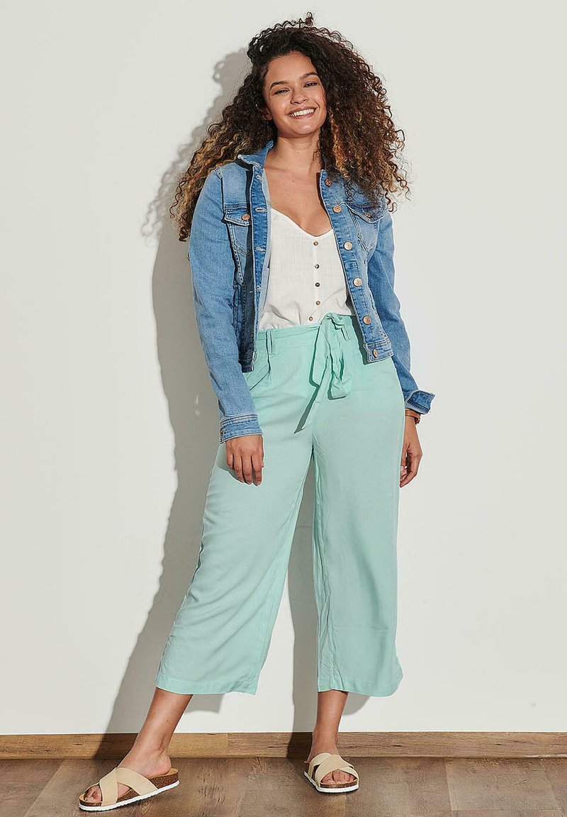 Femeie cu păr creț zâmbind, îmbrăcată cu o jachetă din denim albastră, o bluză albă cu nasturi, pantaloni largi mint verzi și sandale bej fără toc.