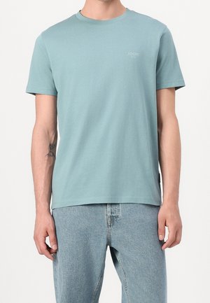 Hombre con camiseta de manga corta azul clara con pequeño logo en el pecho y vaqueros azul claro, de pie frente a un fondo liso.