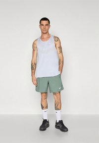 Camiseta sin mangas blanca de malla, pantalones cortos deportivos verdes, zapatillas negras y calcetines blancos. Presenta logos de Nike en la camiseta y los pantalones cortos, con brazos tatuados visibles.