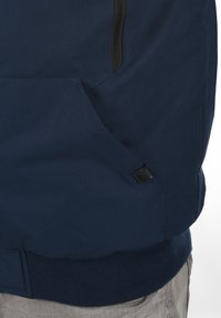 Marineblå jakke med en glat tekstur, som har en lynlåslomme og ribbet kant. Inkluderer et subtilt logo-tag nær lommen.