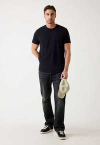 Maglietta nera a girocollo, jeans scuri e sneakers nere. Tiene in mano un berretto da baseball verde chiaro. Outfit semplice e casual su un modello.